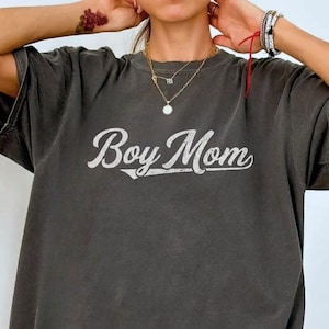 May include: Dark grey t-shirt featuring the text "Boy Mom" in white script. The shirt has a loose fit and short sleeves. A small oval tag at the bottom right says "Comfort Colors Pepper".