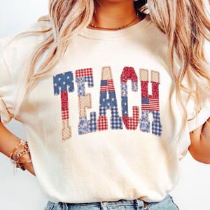 Pode incluir: Uma t-shirt de manga curta na cor marfim com a palavra TEACH impressa em letras grandes de bloco. Cada letra é preenchida com um padrão de retalhos de tecidos vermelhos, brancos e azuis, incluindo estrelas, listras e xadrez vichy.