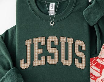 May include: A forest green crewneck sweatshirt featuring the word JESUS in a beige and white checkered varsity-style font across the chest. The garment has long sleeves, a ribbed neckline, and a Gildan brand tag visible at the inner collar.