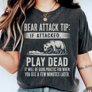 May include: Dark grey heather t-shirt with the text "BEAR ATTACK TIP: IF ATTACKED, PLAY DEAD." The graphic shows a bear and a person lying down. The shirt also says "IT WILL BE GOOD PRACTIC FOR WHEN YOU DIE A FEW MINUTES LATER."