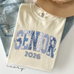 以下が含まれることがあります： アイボリー色のTシャツ。青色のパッチワーク文字で「SENIOR」と書かれ、白いデイジーで装飾され、下に「2026」の年号。薄い色のデニムジーンズ、金のネックレス、ベージュのフェドラハットと組み合わせています。