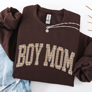 May include: A dark chocolate brown crewneck sweatshirt featuring the text BOY MOM in a beige and brown plaid pattern across the chest. The sweatshirt has long sleeves and a ribbed collar, with a silver-toned chain necklace resting on the neckline.