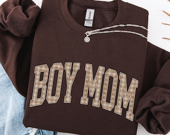 May include: A dark chocolate brown crewneck sweatshirt featuring the text BOY MOM in a beige and brown plaid pattern across the chest. The sweatshirt has long sleeves and a ribbed collar, with a silver-toned chain necklace resting on the neckline.