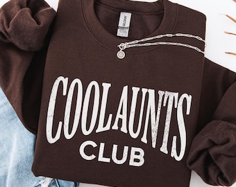 May include: A dark chocolate brown crewneck sweatshirt featuring the white distressed text COOL AUNTS CLUB printed across the chest. The garment has long sleeves and a ribbed collar, paired with a silver chain necklace and blue denim jeans.