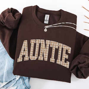 May include: A dark chocolate-colored sweatshirt with the word "AUNTIE" in a plaid design. A silver necklace with a pendant rests on the sweatshirt. The sweatshirt is folded next to a pair of blue jeans.