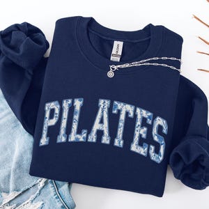 May include: Navy blue Pilates sweatshirt with white floral-patterned lettering. The sweatshirt is folded over a pair of blue jeans. A silver necklace with a small pendant is draped over the sweatshirt. The word "navy" is written in cursive in the bottom right corner.