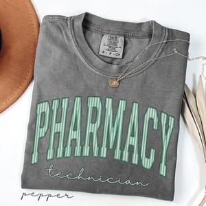 K&ouml;nnte beinhalten: Ein anthrazitfarbenes Kurzarm-T-Shirt mit Rundhalsausschnitt. Das Shirt zeigt das Wort PHARMACY in gro&szlig;en, gebogenen, hellgr&uuml;n gestreiften Blockbuchstaben, darunter steht das Wort technician in einer kleineren, hellgr&uuml;nen Schreibschrift.
