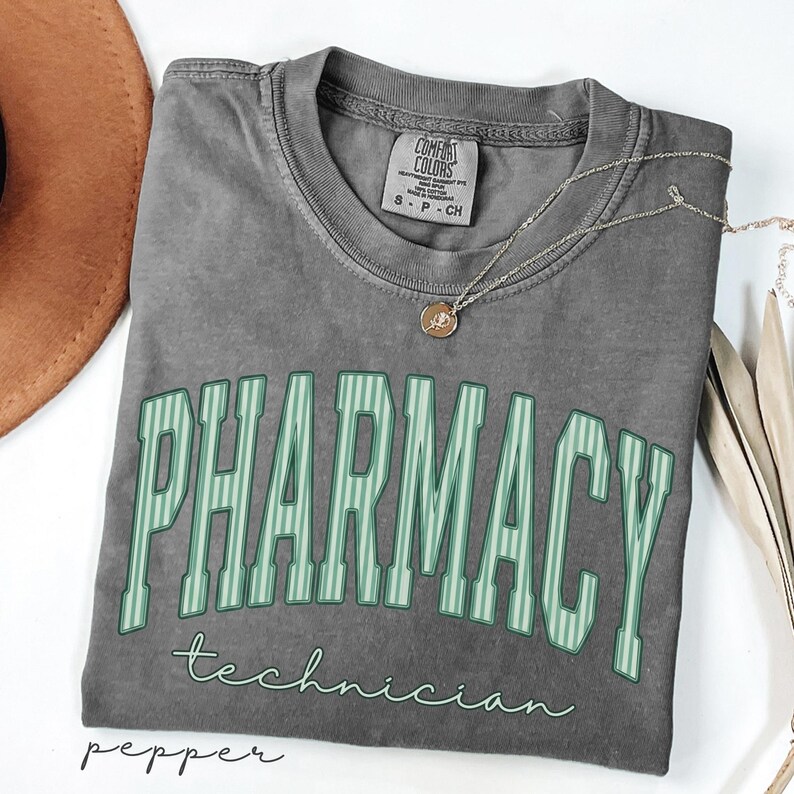K&ouml;nnte beinhalten: Ein anthrazitfarbenes Kurzarm-T-Shirt mit Rundhalsausschnitt. Das Shirt zeigt das Wort PHARMACY in gro&szlig;en, gebogenen, hellgr&uuml;n gestreiften Blockbuchstaben, darunter steht das Wort technician in einer kleineren, hellgr&uuml;nen Schreibschrift.