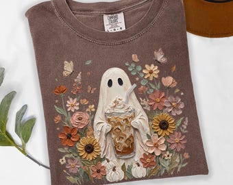May include: A brown short-sleeve t-shirt featuring a graphic of a white ghost holding an iced coffee drink, surrounded by colorful flowers, butterflies, and small white pumpkins. The shirt has a crew neckline and a Comfort Colors brand label.