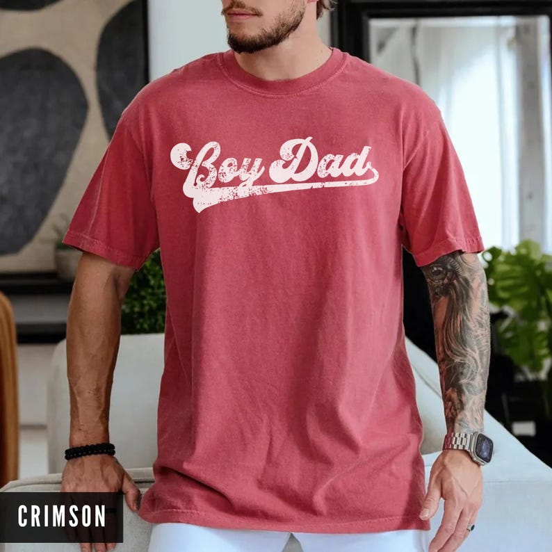 Puede incluir: Camiseta color carmes&iacute; con el texto "Boy Dad" en fuente blanca desgastada. La camiseta de manga corta y cuello redondo la lleva una persona. La palabra "CRIMSON" est&aacute; en la parte inferior de la imagen.