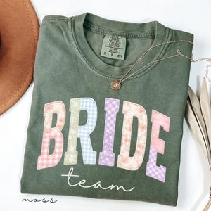 Puede incluir: Una camiseta verde oliva con la palabra "BRIDE" en letras de patchwork, con estampados florales y de cuadros. La palabra "team" está escrita debajo en una fuente de escritura. Un collar de oro con un colgante descansa sobre la camiseta.