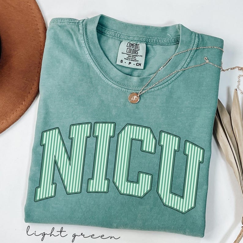 May include: A sage green short-sleeve t-shirt with a crew neckline and a Comfort Colors brand label. The front features the text NICU in large, block-style letters with a vertical light green and white striped pattern. A thin gold-tone necklace is present.