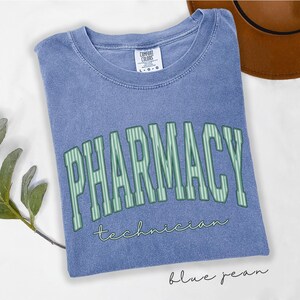K&ouml;nnte beinhalten: Ein blaues T-Shirt mit Rundhalsausschnitt und Vintage-Waschung. Auf der Vorderseite steht das Wort PHARMACY in gro&szlig;en, hellgr&uuml;nen, vertikal gestreiften Blockbuchstaben mit dunkler Umrandung und das Wort technician in kleinerer, gr&uuml;ner Schreibschrift.