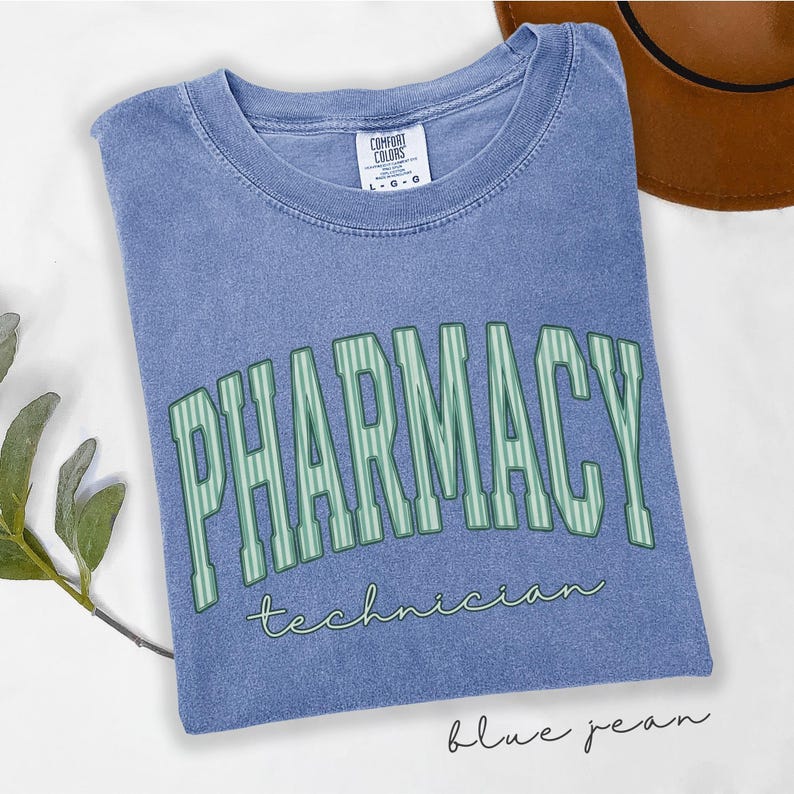 K&ouml;nnte beinhalten: Ein blaues T-Shirt mit Rundhalsausschnitt und Vintage-Waschung. Auf der Vorderseite steht das Wort PHARMACY in gro&szlig;en, hellgr&uuml;nen, vertikal gestreiften Blockbuchstaben mit dunkler Umrandung und das Wort technician in kleinerer, gr&uuml;ner Schreibschrift.