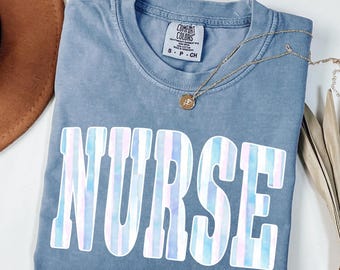 May include: A blue jean colored t-shirt with a crew neckline and a Comfort Colors brand label. The front features the word NURSE in large, block letters with a vertical striped pattern in shades of light blue, pink, and white. A gold necklace is visible.
