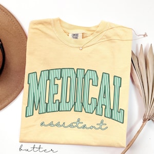 May include: A butter yellow short-sleeve t-shirt with a crew neckline and a Comfort Colors brand label. The front features the text MEDICAL in large green and white striped block letters, with the word assistant in a smaller green cursive font.