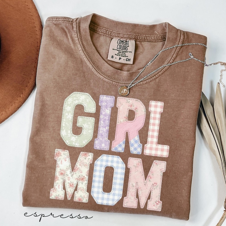 May include: Espresso-colored t-shirt with the words "GIRL MOM" in patchwork letters. The letters feature floral, gingham, and solid pastel patterns. A silver necklace with a pendant rests on the shirt.