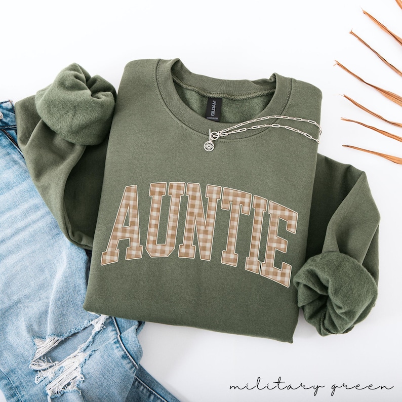 May include: A military green sweatshirt with the word "AUNTIE" in a plaid design across the front. A silver necklace with a pendant is draped on the shirt. The text "military green" is at the bottom of the image.