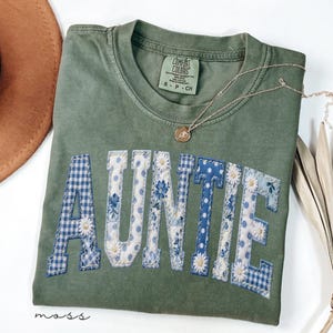 May include: An olive green t-shirt with the word "AUNTIE" in a patchwork design. The letters feature blue gingham, floral, and polka dot patterns. A gold necklace with a pendant rests on the shirt. The shirt is labeled "Comfort Colors".