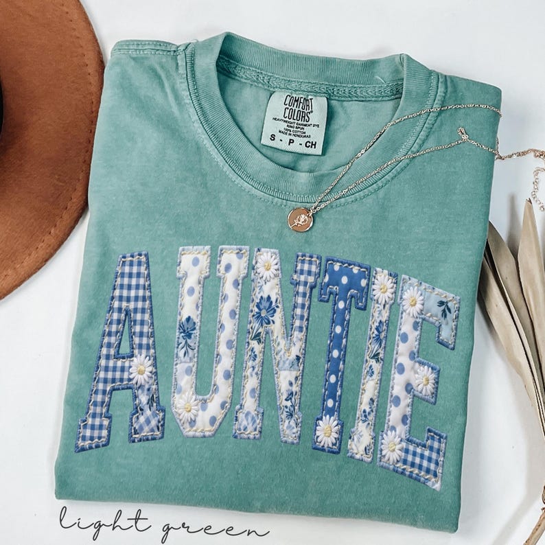 May include: A light green Comfort Colors t-shirt with the word "AUNTIE" appliqued across the front in a collegiate style. The letters feature various blue floral and gingham patterns. A gold necklace with a pendant rests on the shirt.
