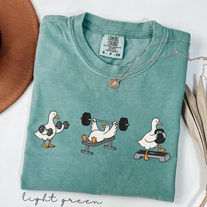 May include: Light green t-shirt featuring a cartoon duck weightlifting design. The graphic shows ducks lifting dumbbells, a barbell, and a kettlebell. The shirt has a crew neck and short sleeves. A silver necklace with a pendant is on the shirt.