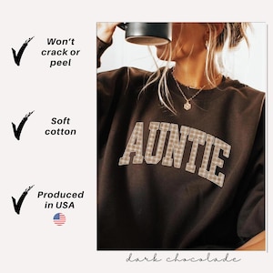 May include: Dark chocolate-colored sweatshirt with the word "AUNTIE" in a plaid design across the front. The sweatshirt is made of soft cotton and produced in the USA. The text "Won't crack or peel" is also visible.