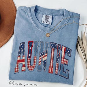 May include: A blue Comfort Colors t-shirt with the word "AUNTIE" in a patriotic design. The letters feature red, white, and blue patterns, including stars and stripes. A gold necklace and a brown hat are also visible.