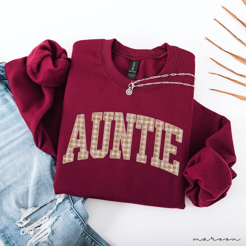 May include: A maroon sweatshirt with the word "AUNTIE" in large, tan plaid letters across the front. A silver necklace with a small pendant rests on the neckline. The sweatshirt is folded on top of a pair of blue jeans.