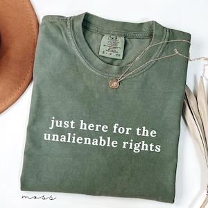 Puede incluir: Una camiseta de manga corta color verde musgo con cuello redondo y el texto en blanco "just here for the unalienable rights" impreso en el frente. La prenda incluye una etiqueta de la marca Comfort Colors en el cuello y un collar dorado fino.