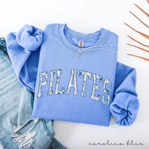 May include: A Carolina blue Pilates sweatshirt with the word "PILATES" in a floral design across the front. A silver necklace with a small pendant rests on the sweatshirt. The sweatshirt is folded on top of a pair of blue jeans.
