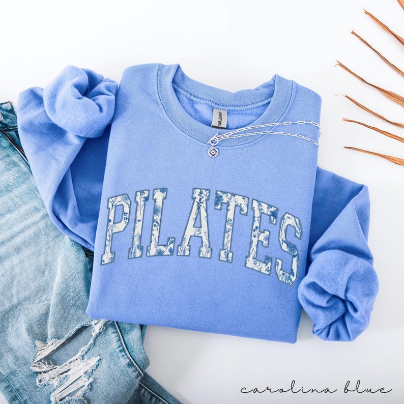 May include: A Carolina blue Pilates sweatshirt with the word "PILATES" in a floral design across the front. A silver necklace with a small pendant rests on the sweatshirt. The sweatshirt is folded on top of a pair of blue jeans.