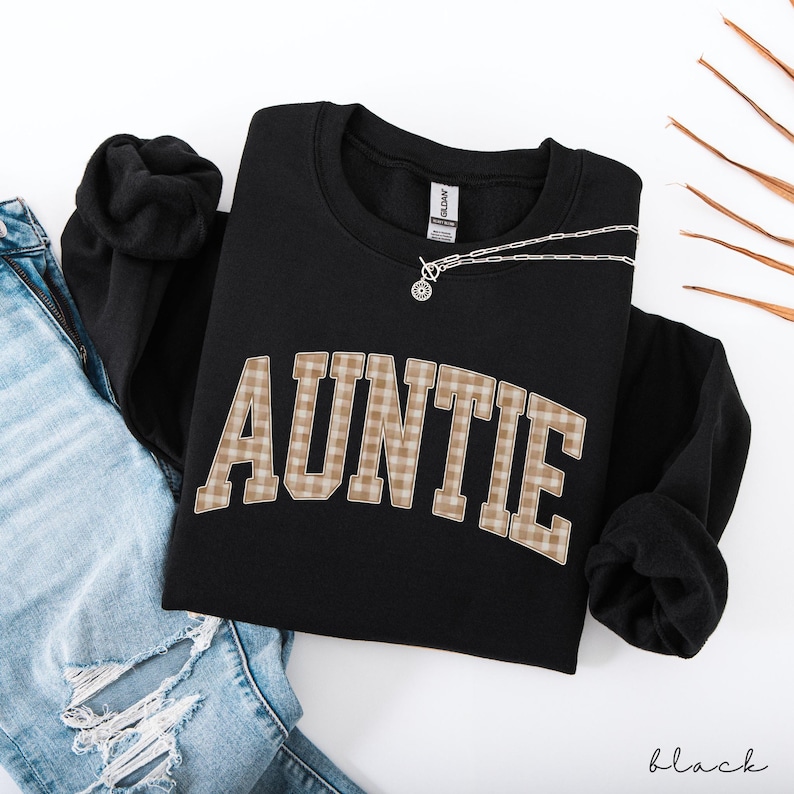 May include: Black sweatshirt with the word "AUNTIE" in a plaid pattern across the front. A silver necklace with a circular pendant rests on the shirt. The word "black" is written in script at the bottom right.