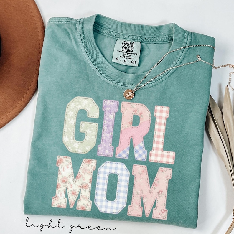 May include: A light green t-shirt with the words "GIRL MOM" in patchwork letters. The letters are in various floral and gingham patterns. A gold necklace with a pendant rests on the shirt. The words "light green" are written in cursive at the bottom.