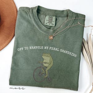 May include: A moss green t-shirt with the text "OFF TO WRANGLE MY FERAL GRANDKIDS" and a cartoon frog riding a bicycle. The shirt has a Comfort Colors tag and a small gold necklace. The shirt is folded on a white surface.