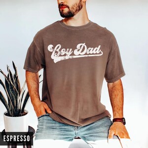 Puede incluir: Camiseta color espresso con la frase "Boy Dad" en fuente blanca desgastada. La camiseta tiene mangas cortas y cuello redondo. El modelo lleva vaqueros azules y un reloj marr&oacute;n.