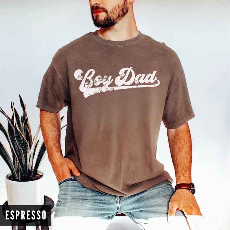 Puede incluir: Camiseta color espresso con la frase "Boy Dad" en fuente blanca desgastada. La camiseta tiene mangas cortas y cuello redondo. El modelo lleva vaqueros azules y un reloj marr&oacute;n.