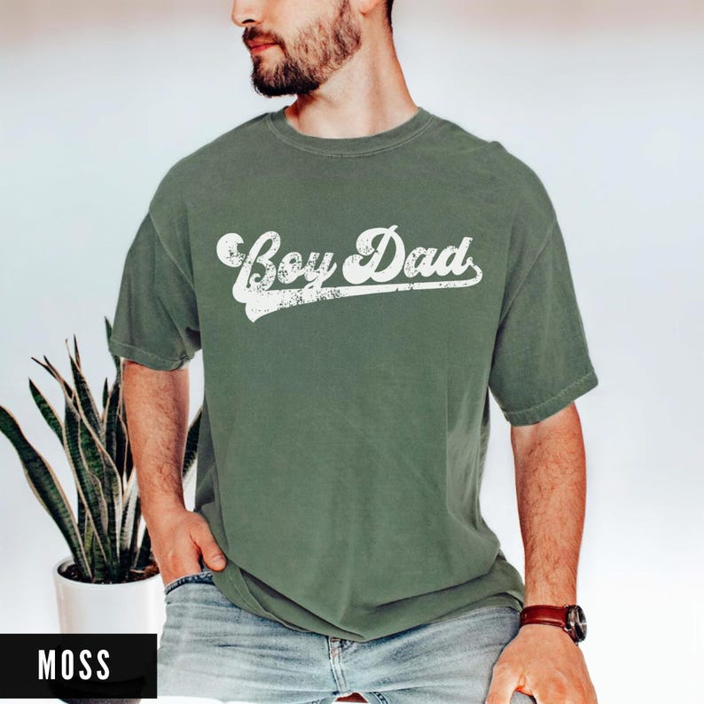 Puede incluir: Camiseta verde musgo con el texto "Boy Dad" en fuente blanca desgastada. La camiseta tiene mangas cortas y cuello redondo. El modelo lleva un reloj marr&oacute;n.