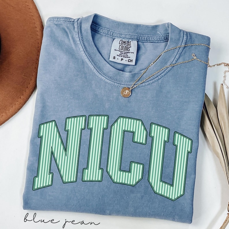 May include: A blue jean colored short-sleeve t-shirt with a crew neckline. The shirt features the text NICU in large, green and white vertical striped block letters across the chest. A Comfort Colors brand label is visible at the inner neckline.