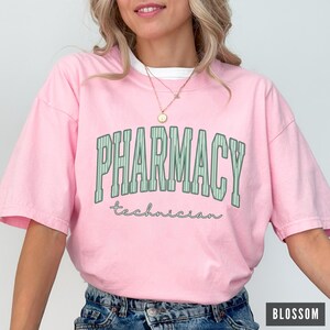 K&ouml;nnte beinhalten: Ein hellrosa T-Shirt mit kurzen &Auml;rmeln, das das Wort PHARMACY in gro&szlig;en, gr&uuml;nen, vertikal gestreiften Blockbuchstaben im College-Stil zeigt, mit dem Wort technician darunter in einer kleineren, kursiven, dunkelgr&uuml;nen Schrift.