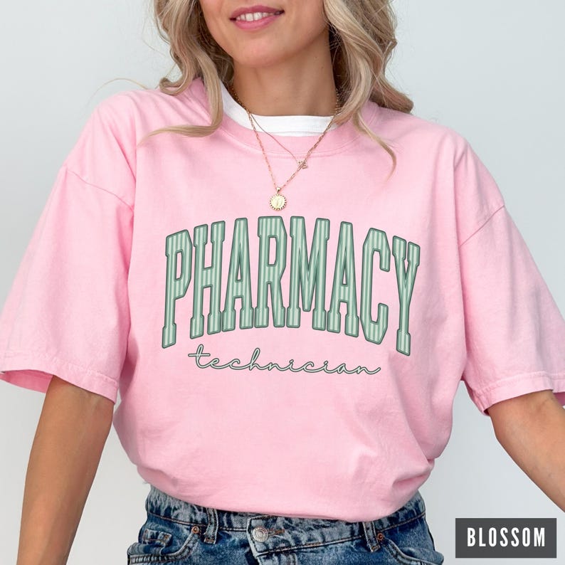 K&ouml;nnte beinhalten: Ein hellrosa T-Shirt mit kurzen &Auml;rmeln, das das Wort PHARMACY in gro&szlig;en, gr&uuml;nen, vertikal gestreiften Blockbuchstaben im College-Stil zeigt, mit dem Wort technician darunter in einer kleineren, kursiven, dunkelgr&uuml;nen Schrift.