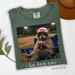 Può includere: T-shirt verde Comfort Colors con una grafica di un procione con occhiali da sole e un cappello "Raising Ballers", che beve da una tazza, seduto su una sedia blu a una partita di baseball. Il testo "i live here now" è sotto.