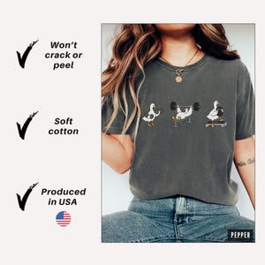 May include: Dark grey t-shirt with three cartoon ducks lifting weights. The shirt has the text "Won't crack or peel", "Soft cotton", and "Produced in USA". The shirt is made of soft cotton.