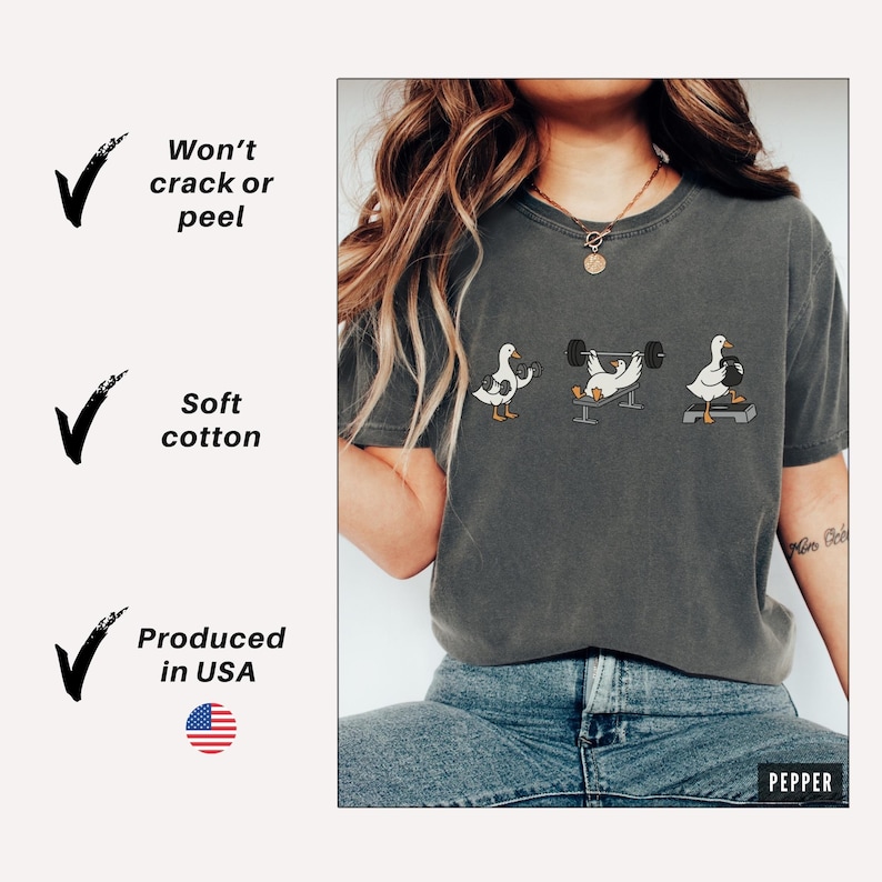 May include: Dark grey t-shirt with three cartoon ducks lifting weights. The shirt has the text "Won't crack or peel", "Soft cotton", and "Produced in USA". The shirt is made of soft cotton.