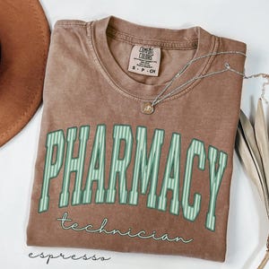 K&ouml;nnte beinhalten: Ein braunes kurz&auml;rmeliges T-Shirt mit Rundhalsausschnitt. Das Shirt zeigt das Wort PHARMACY in gro&szlig;en, hellgr&uuml;n gestreiften Blockbuchstaben, darunter steht das Wort technician in einer kleineren, kursiven wei&szlig;en Schrift. Eine Halskette ist zu sehen.