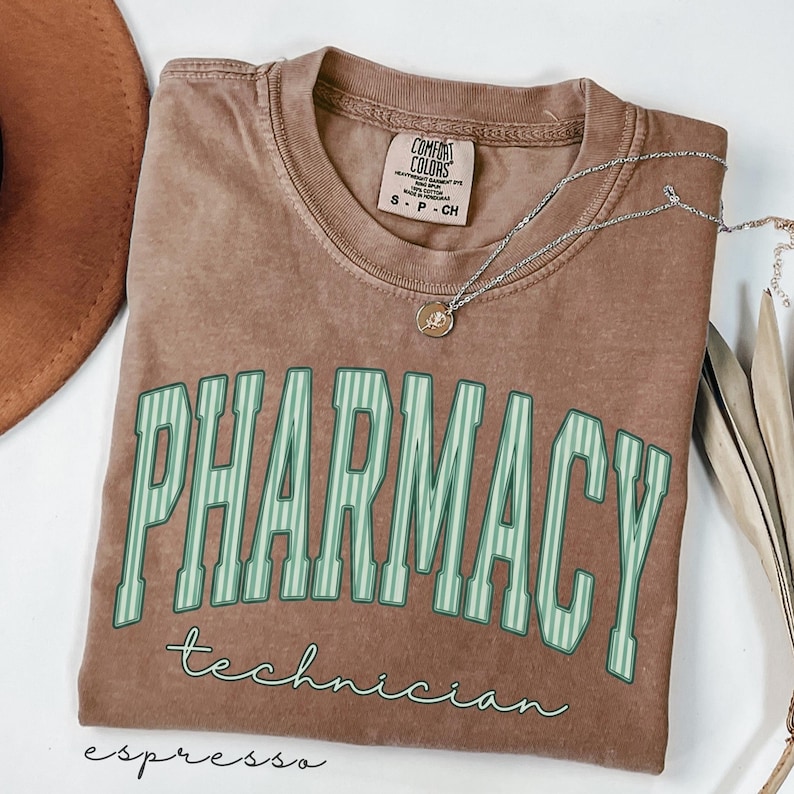 K&ouml;nnte beinhalten: Ein braunes kurz&auml;rmeliges T-Shirt mit Rundhalsausschnitt. Das Shirt zeigt das Wort PHARMACY in gro&szlig;en, hellgr&uuml;n gestreiften Blockbuchstaben, darunter steht das Wort technician in einer kleineren, kursiven wei&szlig;en Schrift. Eine Halskette ist zu sehen.