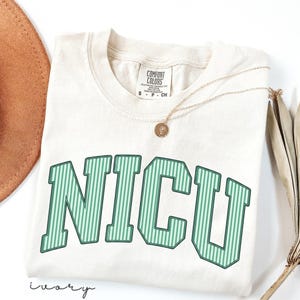 May include: An ivory short-sleeve t-shirt with a crew neckline and a Comfort Colors brand label. The front features the text NICU in large, block-style letters with a green and white vertical striped pattern and a dark green outline.