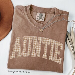May include: Espresso-colored t-shirt with the word "AUNTIE" in a plaid pattern. The shirt has a crew neck and short sleeves. A silver necklace with a small pendant is draped across the shirt. The shirt is a Comfort Colors brand.