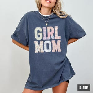 May include: A denim blue t-shirt with the words "GIRL MOM" in pastel patchwork letters. The shirt has a crew neck and short sleeves. The word "DENIM" is in a white box in the lower right corner.