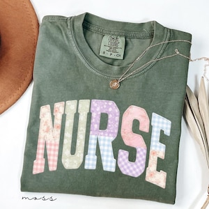 Puede incluir: Una camiseta verde oliva con la palabra "NURSE" en letras de patchwork. Las letras presentan patrones florales, de cuadros y pastel. Un collar dorado con un pequeño colgante descansa sobre la camiseta. Un sombrero marrón es visible en la esquina superior izquierda.