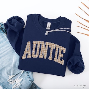 May include: Navy blue sweatshirt with the word "AUNTIE" in a plaid pattern across the front. A silver necklace with a pendant is draped over the sweatshirt. The sweatshirt is folded next to a pair of blue jeans.